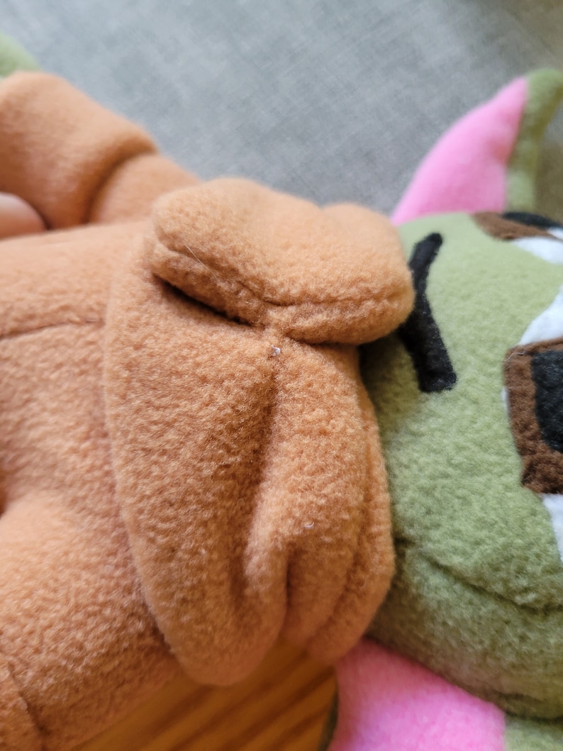 May include: A plush toy with brown, green, and pink fabric. The toy has a brown hooded head and a green body with a pink ear.