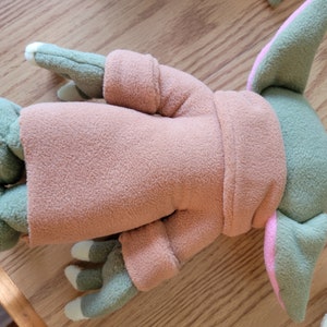 May include: A plush toy of a green and pink character wearing a brown hooded robe. The toy is lying on its back with its arms and legs outstretched.