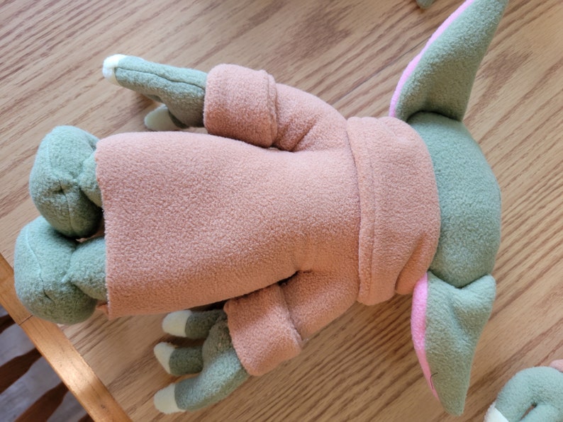 May include: A plush toy of a green and pink character wearing a brown hooded robe. The toy is lying on its back with its arms and legs outstretched.