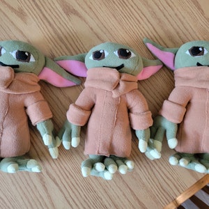 May include: Three plush toys of a popular fictional character. The toys are green and brown with large eyes and pink ears. They are all wearing brown coats.