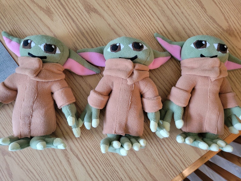 May include: Three plush toys of a popular fictional character. The toys are green and brown with large eyes and pink ears. They are all wearing brown coats.