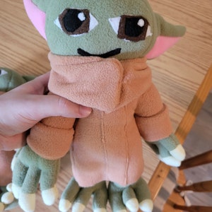 May include: A green plush toy with large brown eyes and a pink nose. The toy is wearing a brown fleece jacket and has four green fingers on each hand.