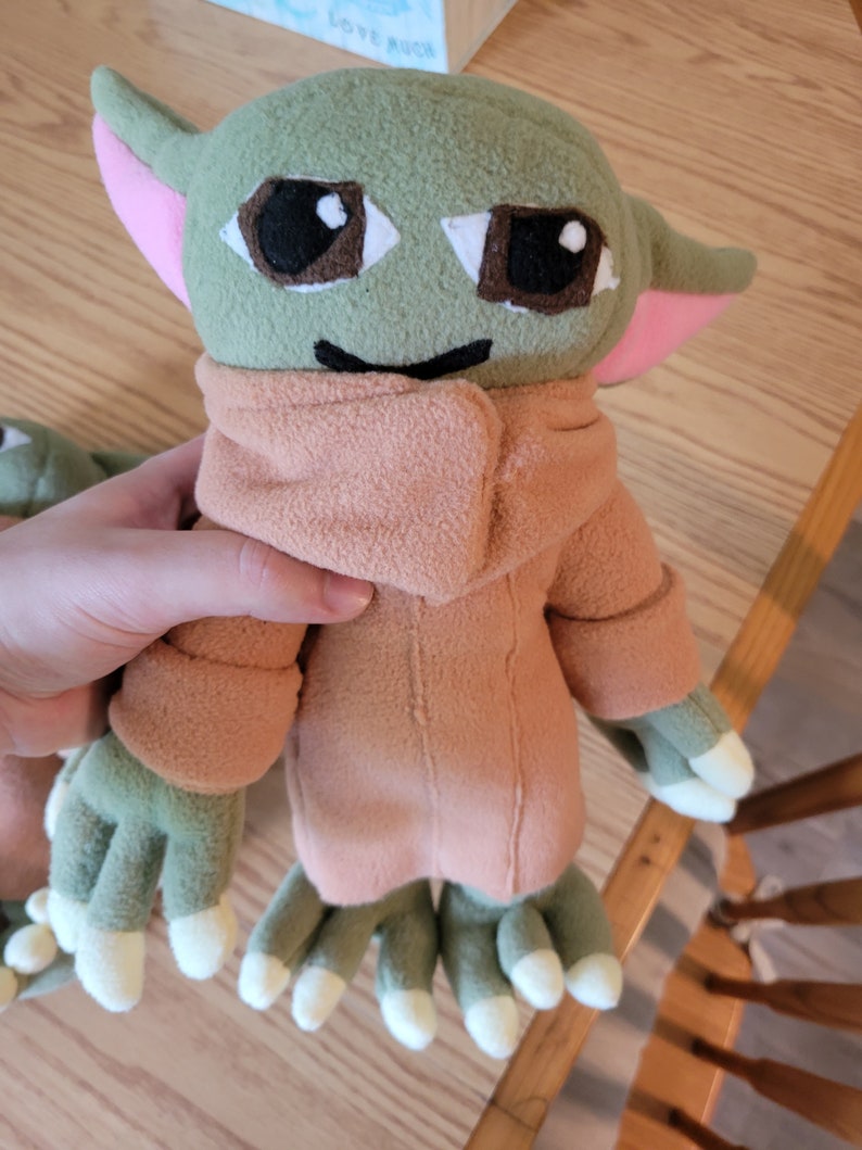May include: A green plush toy with large brown eyes and a pink nose. The toy is wearing a brown fleece jacket and has four green fingers on each hand.