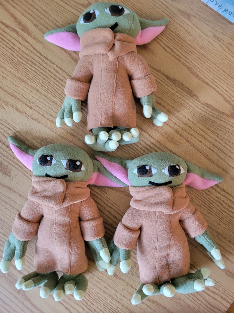 May include: Three plush toys of a popular fictional character. The toys are green, brown, and pink. The toys are all wearing brown coats. The toys are all smiling.