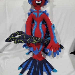 May include: A handmade plush toy of a fantastical creature. The toy is primarily red with blue accents, including the head, arms, and tail. The creature has a smiling face and a detailed, patterned design. The toy is made of soft fabric.