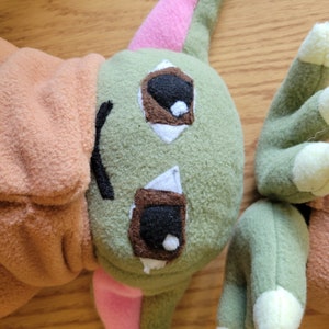 May include: A green plush toy with pink ears and brown eyes. The toy is made of soft, fuzzy fabric and has a friendly expression.