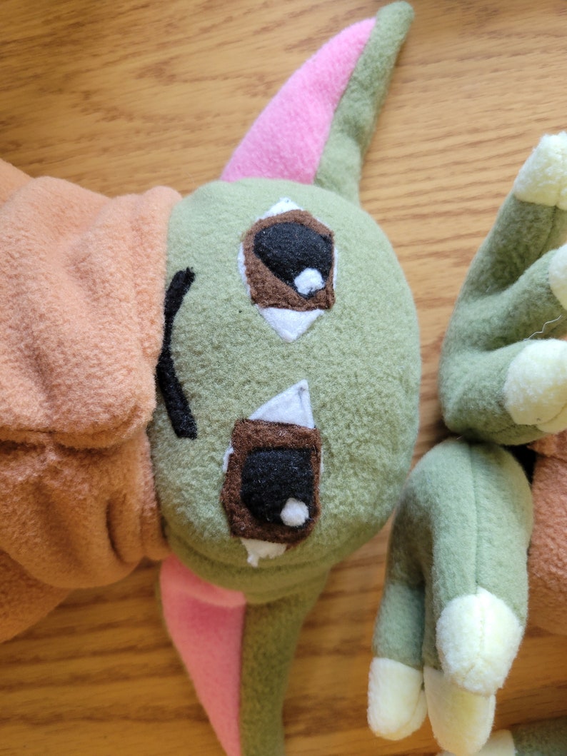May include: A green plush toy with pink ears and brown eyes. The toy is made of soft, fuzzy fabric and has a friendly expression.
