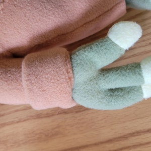 May include: Close-up of a plush toy's arm, featuring a light brown fleece sleeve and a green hand with three cream-colored fingers.