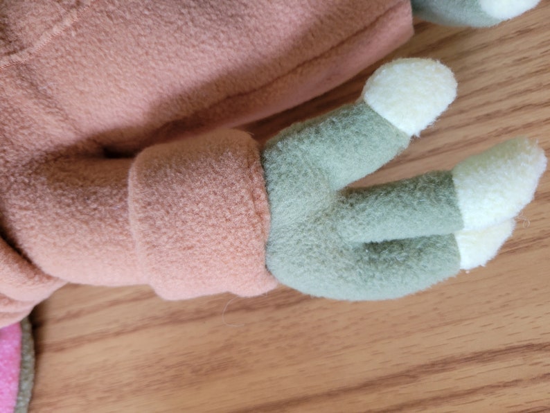 May include: Close-up of a plush toy's arm, featuring a light brown fleece sleeve and a green hand with three cream-colored fingers.