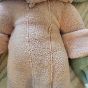 May include: A plush toy with a brown hooded jacket and green and pink accents. The jacket has a seam down the front.
