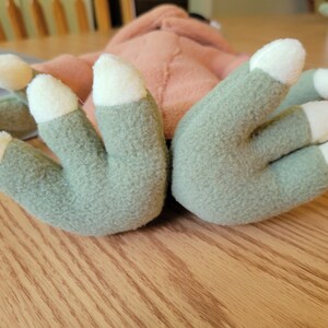 May include: Close-up of a plush toy with green hands and white claws. The toy is lying on a wooden surface.