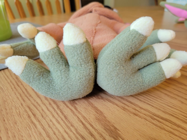 May include: Close-up of a plush toy with green hands and white claws. The toy is lying on a wooden surface.