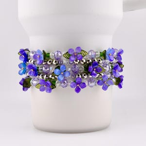 Pode incluir: Uma caneca de cerâmica branca é adornada com uma pulseira com contas de flores roxas e azuis. A pulseira envolve a caneca, mostrando o design floral e as contas prateadas. As flores têm folhas verdes.