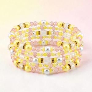Op de afbeelding: Een armband met meerdere strengen met gele, roze, witte en zilveren kralen. De armband heeft een spiraalvormig ontwerp met een mix van ronde en facetgeslepen kralen. De kralen zijn geregen met een flexibele draad, waardoor een gelaagd effect ontstaat.