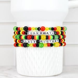 May include: Four beaded bracelets with the words "CELEBRATE BLACK HISTORY" in white letter beads. The bracelets feature red, yellow, green, and black beads. The bracelets are stacked on a white ceramic mug.