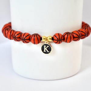Peut inclure: Un bracelet sur le thème du basketball avec des perles orange et des lignes noires, imitant des ballons de basket. Il comprend un charm doré avec la lettre "K" sur fond noir. Le bracelet est présenté sur fond blanc.