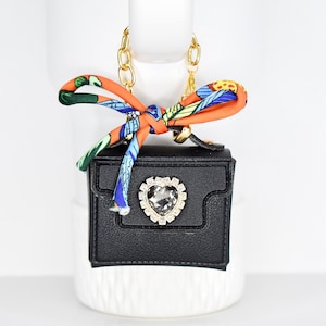 May include: A small, black handbag with a gold chain strap and a decorative bow. The bow is orange with a colorful pattern. The bag features a heart-shaped rhinestone embellishment. The bag is approximately 3 inches wide.