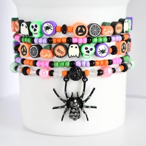 以下が含まれることがあります： オレンジ、黒、緑、ピンク、紫色のハロウィーンをテーマにしたビーズブレスレット。ブレスレットには、幽霊、スカル、カボチャ、クモなど、さまざまなハロウィーンをテーマにしたチャームが付いています。黒いクモのチャームが下のブレスレットからぶら下がっています。