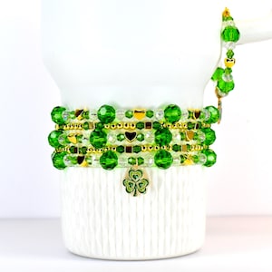 May include: A stack of green beaded bracelets with gold accents and heart charms. A single bracelet with a gold chain and green beads is on the side. A gold shamrock charm hangs from the bottom bracelet. The bracelets are displayed on a white surface.