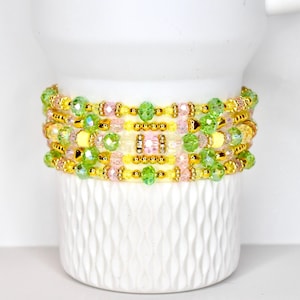 May include: A stack of beaded bracelets in shades of yellow, pink, and green. The bracelets are adorned with gold-colored beads and clear, faceted beads. The bracelets are displayed on a white, textured surface.