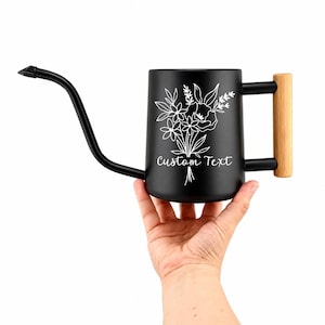 Puede incluir: Una regadera de color negro mate con un pico largo y delgado y un mango de madera clara. El lateral de la regadera presenta una ilustración de líneas blancas de un ramo de flores sobre el texto Custom Text en una fuente cursiva.