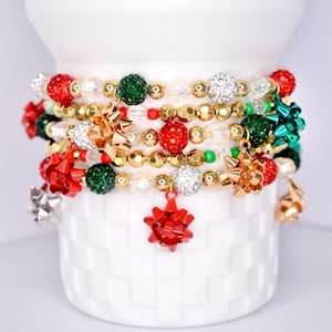 Könnte beinhalten: Eine Sammlung von Armbändern mit roten, grünen, goldenen und silbernen Perlen. Die Armbänder weisen verschiedene Perlen Größen und Texturen auf, darunter funkelnde Kristallperlen und kleine Schleifenanhänger. Präsentiert auf einer weißen, strukturierten Oberfläche.