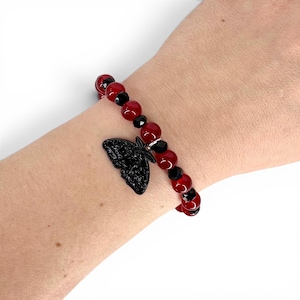 Op de afbeelding: Een kralenarmband met afwisselend gladde rode bolvormige kralen en kleinere gefacetteerde zwarte kralen. Een zwarte bedel in de vorm van een nachtvlinder hangt in het midden. De kralen zijn geregen aan een elastisch koord voor een ronde vorm.