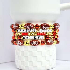 Op de afbeelding: Gestapelde armbanden met kralen in de vorm van voetballen, gouden en rode kralen, die "KANSAS CITY CHIEFS" spellen met witte letterkralen. De armbanden zijn gestapeld op een witte keramische mok.