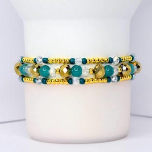 Może przedstawiać: Trzyrzędowa bransoletka z koralikami w kolorze złotym, turkusowym i przezroczystym. Bransoletka ma złote przekładki i fasetowane złote koraliki. Turkusowe koraliki są jednolite, a przezroczyste koraliki są małe i okrągłe.