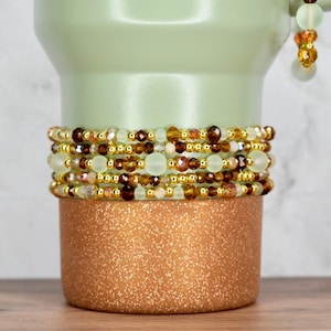 May include: A stack of beaded bracelets in shades of brown, gold, and cream, with a mix of small and large beads. The bracelets are displayed on a copper-colored, glittery cylindrical base, with a light green background.