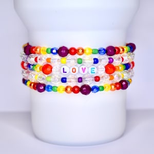 Può includere: Una pila di braccialetti di perline colorate. Ogni braccialetto presenta un arcobaleno di perline, tra cui trasparenti, viola, rosse, arancioni, gialle, verdi e blu. Un braccialetto riporta la parola "LOVE" con perline di lettere bianche. I braccialetti sono esposti su un oggetto cilindrico bianco.