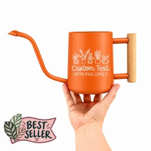 May include: An orange metal watering can with a long, thin spout and a light-colored wooden handle. The side features white line art of various potted plants above the text Custom Text and OPTIONAL LINE 2. A Best Seller graphic is visible.