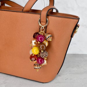 Può includere: Una borsa marrone con un anello in metallo color oro che tiene un gruppo di perline decorative. Le perline sono di vari colori, tra cui giallo, marrone, bordeaux e perla, con alcuni dettagli glitterati. La borsa è di colore marrone chiaro uniforme.