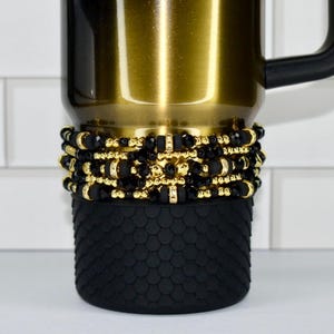 May include: A gold-colored travel mug with a black handle. The mug is adorned with multiple layers of black and gold beaded accents. The bottom of the mug features a textured black rubber sleeve.