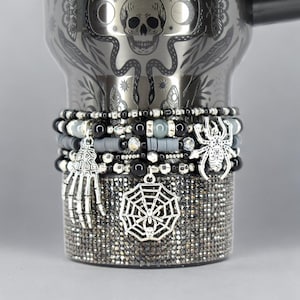 Peut inclure: Un gobelet en acier inoxydable avec un motif de crâne et de serpent, orné de bracelets de perles. Les bracelets comportent des breloques comme une araignée, une main de squelette et une toile d'araignée. Le bas du gobelet est recouvert de strass scintillants.