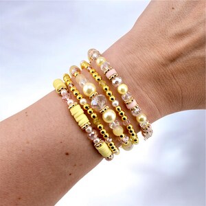May include: A set of five stacked bracelets featuring a combination of yellow clay disc beads, gold-toned metallic spheres, clear faceted crystal beads, and smooth cream-colored pearls. The bracelets are arranged in a layered design on a wrist.
