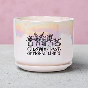 May include: A pearlescent white ceramic planter with a matching saucer. The planter features a black graphic of potted plants and the text "Custom Text Optional Line 2". The planter is designed for indoor use.