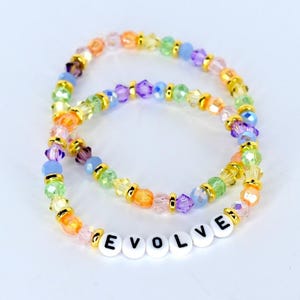 May include: Two beaded bracelets with colourful beads in shades of orange, yellow, green, blue, and purple. One bracelet spells out the word "EVOLVE" with white letter beads. Gold-coloured spacers separate the beads.