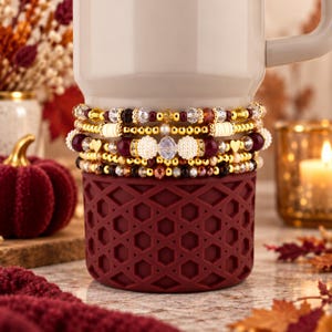 Puede incluir: Una pila de cinco pulseras de cuentas envueltas alrededor de una taza blanca con una funda de silicona color granate. Las pulseras presentan una mezcla de cuentas doradas, cristales facetados transparentes, cuentas granates y pequeños dijes de corazón.