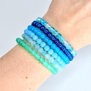 May include: A collection of six beaded bracelets in shades of blue and green. The bracelets are stacked on a wrist, showcasing a gradient of colors from light green to deep blue. The beads are round and vary in size.