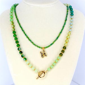 Könnte beinhalten: Halskette mit zwei Strängen aus grünen Perlen und einem goldfarbenen Initialenanhänger. Die Halskette hat einen Knebelverschluss und eine Vielzahl von Grüntönen. Die Perlen sind mit goldfarbenen Akzenten durchsetzt.