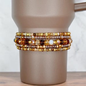 Könnte beinhalten: Eine Sammlung von Armbändern mit Perlen in Gold-, Braun- und Bernsteintönen umschließt einen braunen Becher. Die Armbänder weisen eine Vielzahl von Perlengrößen und -texturen auf, wodurch ein geschichteter Effekt entsteht. Der Becher hat einen Griff.