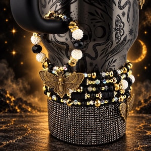 Könnte beinhalten: Ein Stapel aus Perlenarmbändern mit schwarzen und goldenen facettierten Perlen, weißen texturierten Kugeln und einem zentralen antikgoldfarbenen Mottenanhänger mit Totenkopfdetail auf einem schwarzen Becher mit Strass-Sockel.