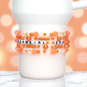 May include: A white mug adorned with multiple beaded bracelets in shades of orange and clear. The bracelets spell out "SHOWGIRL ERA" with white letter beads. The bracelets are wrapped around the mug.