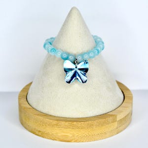 May include: A light blue beaded bracelet with a butterfly charm. The butterfly charm has a blue and white design. The bracelet is displayed on a white cone-shaped stand with a wooden base. The bracelet is a piece of jewelry.