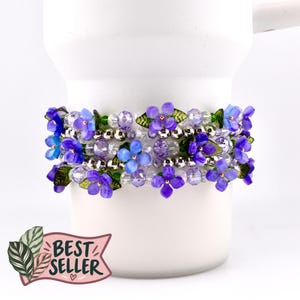 May include: A multi-strand beaded bracelet featuring purple and blue flower-shaped beads, clear faceted crystal beads, metallic silver spherical beads, and small green leaf-shaped accents. A pink banner graphic with the text BEST SELLER is included.