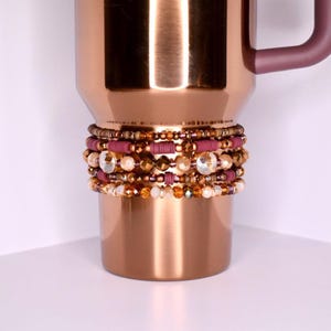 May include: A copper-coloured travel mug with a dark purple handle. The mug is decorated with multiple beaded bracelets in shades of gold, purple, and clear crystals. The bracelets are wrapped around the base of the mug, adding a decorative touch.