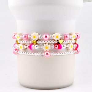 May include: Four beaded bracelets with pink, white, and gold accents. The bracelets feature pearls, daisy-shaped beads, and heart-shaped charms. The bracelets are stacked on a white cylindrical object.