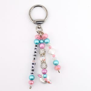 May include: A silver-toned metal keyring featuring three dangling strands of beads. One strand displays white square letter beads spelling BEACH BABE. The design includes pink, blue, and clear crystal-encrusted beads, plus a small white seashell charm.