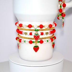 May include: A selection of beaded bracelets in red, gold, and clear beads, with green and gold charms. The bracelets feature strawberry charms and are displayed on a white surface.